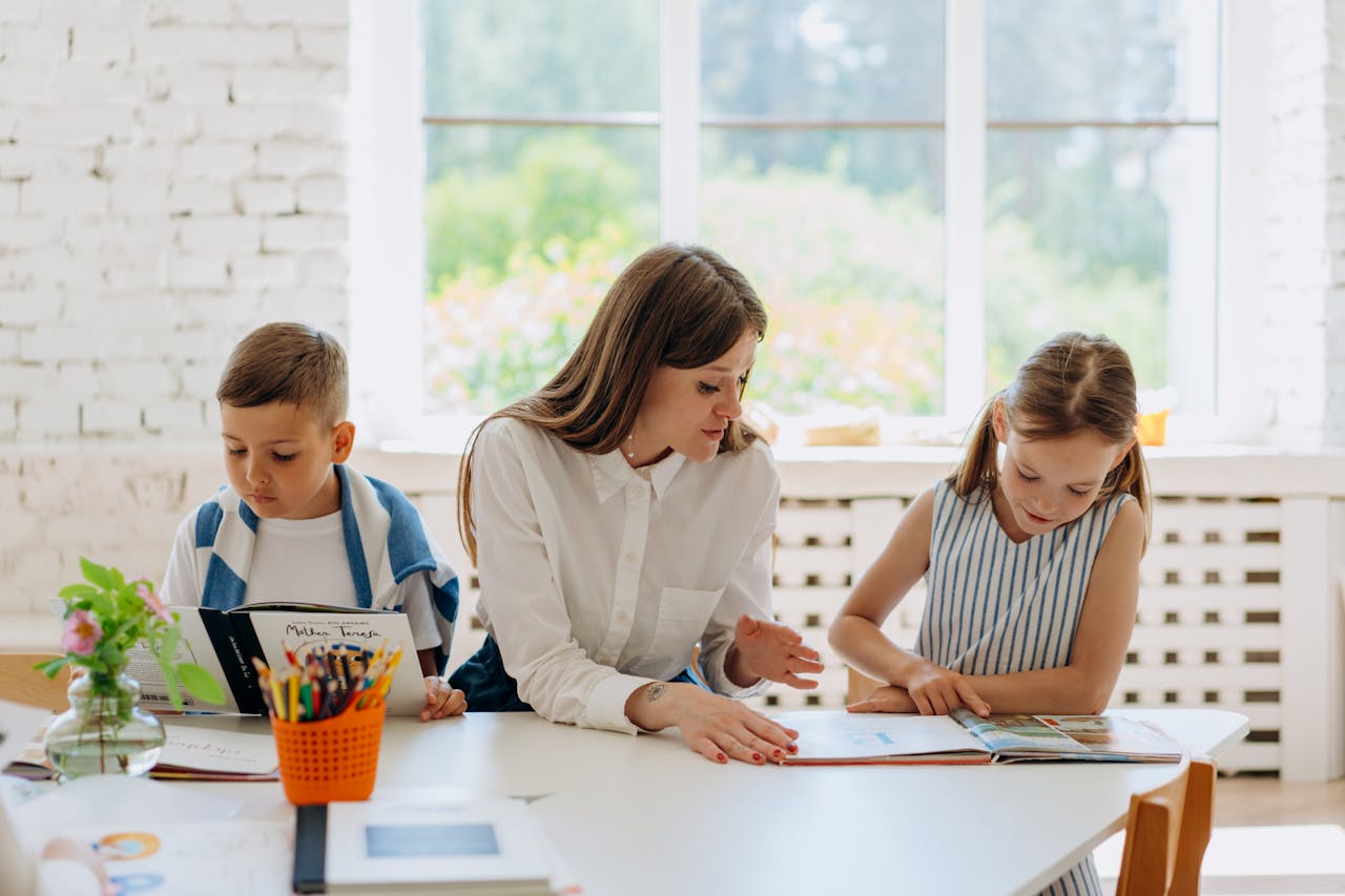 A female teacher helps two children learn by reading together in a bright classroom.