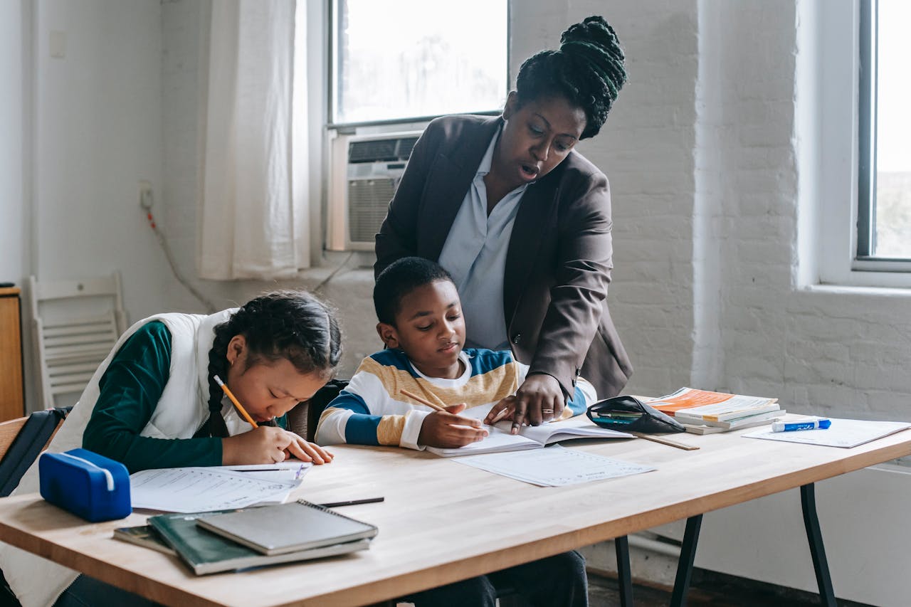 Focused African American female teacher in formal clothes standing near school desk with diverse pupils and pointing at schoolboys copybook